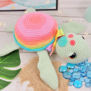 Puede incluir: Un juguete de tortuga marina de crochet con una concha de arco&iacute;ris, un cuerpo verde y ojos azules. La tortuga est&aacute; tumbada sobre una superficie blanca con arena y piedras de vidrio azules.