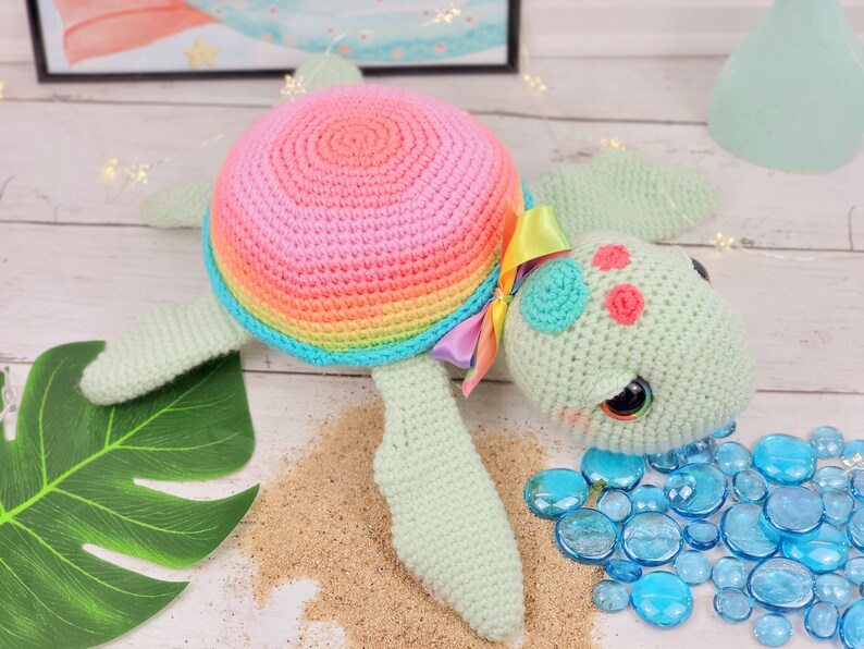 Puede incluir: Un juguete de tortuga marina de crochet con una concha de arco&iacute;ris, un cuerpo verde y ojos azules. La tortuga est&aacute; tumbada sobre una superficie blanca con arena y piedras de vidrio azules.