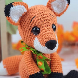 May include: A crocheted orange fox with black eyes, a white nose, and black feet. The fox is wearing a green ribbon with orange leaves.