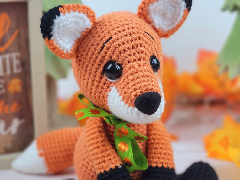 May include: A crocheted orange fox with black eyes, a white nose, and black feet. The fox is wearing a green ribbon with orange leaves.