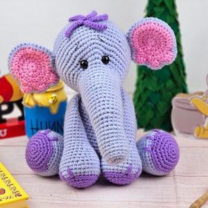 May include: A crocheted purple and pink elephant toy with a purple flower on its head. The elephant is sitting on a wooden surface with a book and a honey pot in the background.