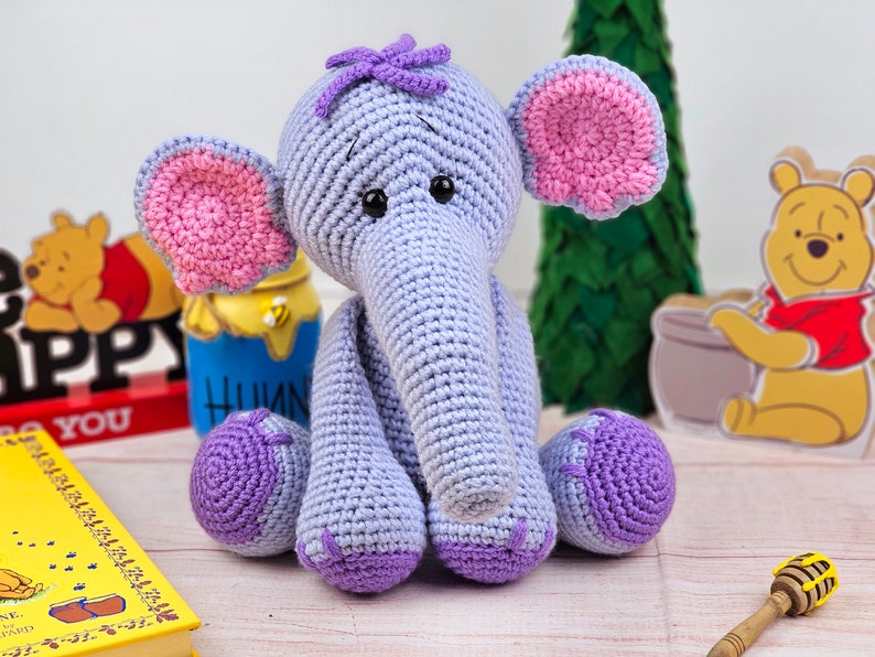 May include: A crocheted purple and pink elephant toy with a purple flower on its head. The elephant is sitting on a wooden surface with a book and a honey pot in the background.