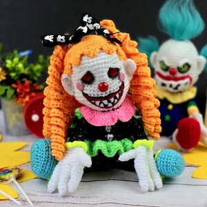 May include: Two crocheted clown dolls, one with orange hair and a black and white bow, and the other with blue hair. Both dolls have a scary expression and are holding lollipops.