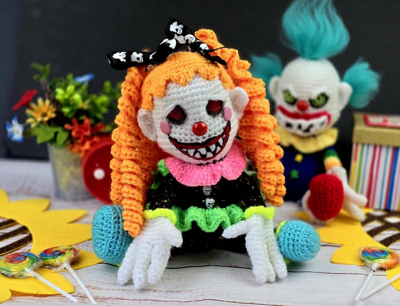 May include: Two crocheted clown dolls, one with orange hair and a black and white bow, and the other with blue hair. Both dolls have a scary expression and are holding lollipops.