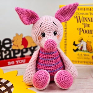 May include: A pink crocheted pig with a striped shirt, sitting on a wooden surface. The pig has a pink nose and black eyes. The pig is surrounded by a sunflower, a book, and a sign that says "Be Happy Do You".