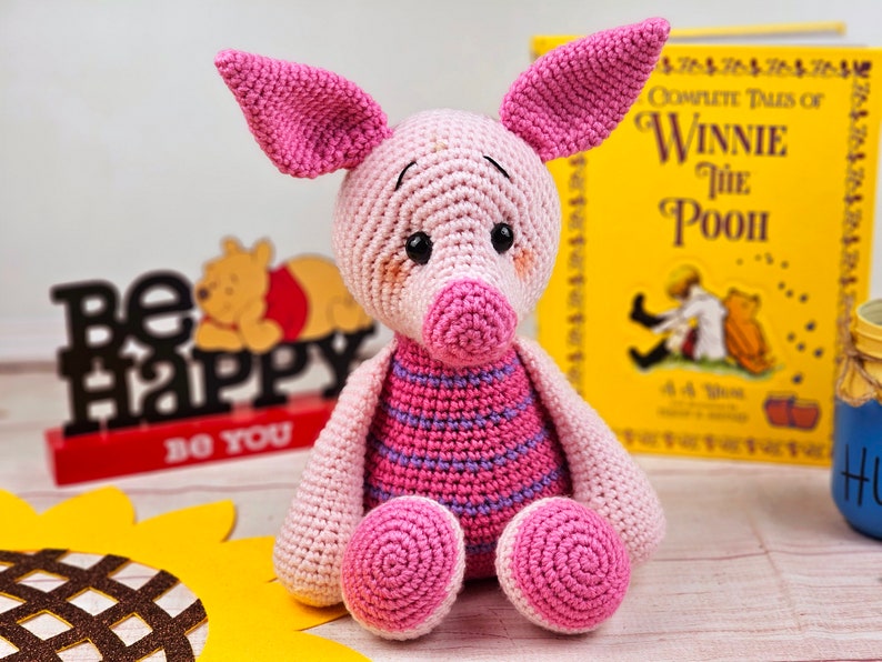 May include: A pink crocheted pig with a striped shirt, sitting on a wooden surface. The pig has a pink nose and black eyes. The pig is surrounded by a sunflower, a book, and a sign that says "Be Happy Do You".