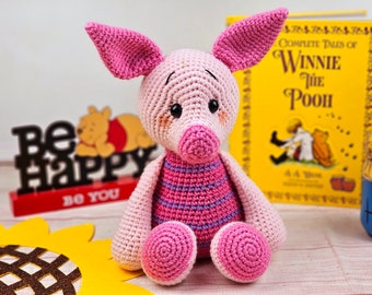 May include: A pink crocheted pig with a striped shirt, sitting on a wooden surface. The pig has a pink nose and black eyes. The pig is surrounded by a sunflower, a book, and a sign that says "Be Happy Do You".
