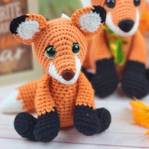 May include: Two crocheted fox plushies, one orange and white with black accents, the other orange with black and white accents. Both have green yarn accents around their necks.