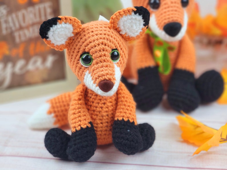 May include: Two crocheted fox plushies, one orange and white with black accents, the other orange with black and white accents. Both have green yarn accents around their necks.