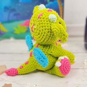 Puede incluir: Un drag&oacute;n de crochet verde con detalles en rosa y azul. El drag&oacute;n est&aacute; sentado sobre una superficie blanca y tiene un ala azul.
