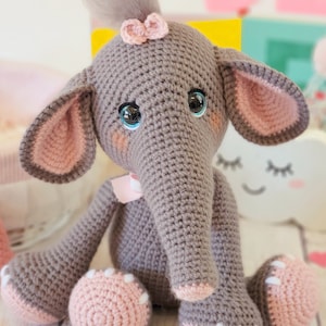 May include: A gray crocheted elephant toy with pink ears, a pink bow, and blue eyes. The elephant is sitting on a white surface.