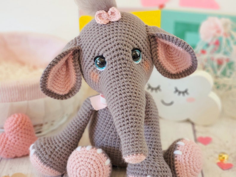 May include: A gray crocheted elephant toy with pink ears, a pink bow, and blue eyes. The elephant is sitting on a white surface.