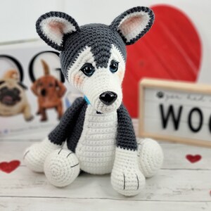 May include: A crocheted husky dog toy with blue eyes, a black nose, and a light blue collar. The toy is sitting on a white wooden surface with red hearts scattered around it. The background includes a book with the word "DOGG" on the cover and a sign that says "You had me at WOOF".