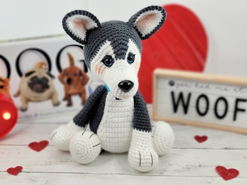 May include: A crocheted husky dog toy with blue eyes, a black nose, and a light blue collar. The toy is sitting on a white wooden surface with red hearts scattered around it. The background includes a book with the word "DOGG" on the cover and a sign that says "You had me at WOOF".