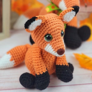 May include: A crocheted orange fox with black and white accents. The fox has large green eyes and a white nose. The fox is sitting on a white surface with fall leaves.