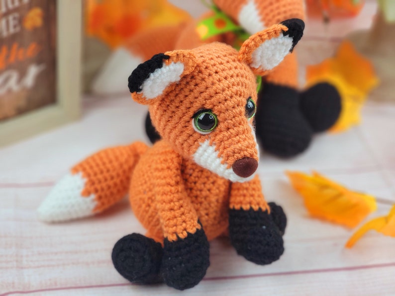 May include: A crocheted orange fox with black and white accents. The fox has large green eyes and a white nose. The fox is sitting on a white surface with fall leaves.