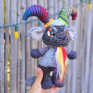 Puede incluir: Un juguete de crochet gris con cuernos de hilo arco&iacute;ris y una cola de hilo arco&iacute;ris. El juguete tiene ojos negros grandes y una peque&ntilde;a nariz negra.