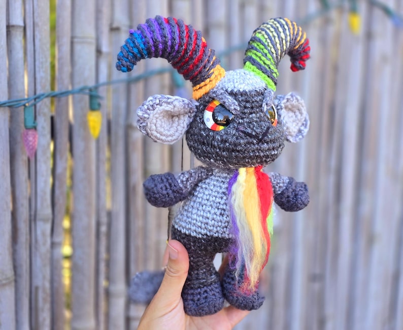 Puede incluir: Un juguete de crochet gris con cuernos de hilo arco&iacute;ris y una cola de hilo arco&iacute;ris. El juguete tiene ojos negros grandes y una peque&ntilde;a nariz negra.