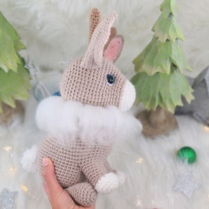 May include: A crocheted bunny with a fluffy white collar, light brown body, and white accents. The bunny has blue eyes and pink inner ears. The background includes green Christmas trees and a star.