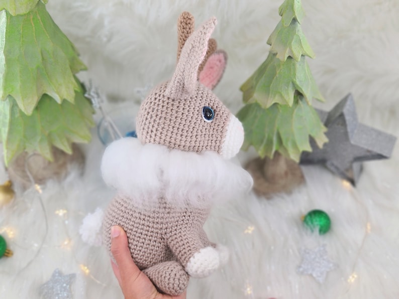 May include: A crocheted bunny with a fluffy white collar, light brown body, and white accents. The bunny has blue eyes and pink inner ears. The background includes green Christmas trees and a star.