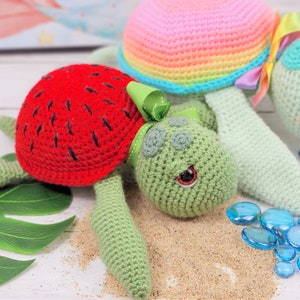 Puede incluir: Dos juguetes de tortuga de crochet, uno con una c&aacute;scara de sand&iacute;a roja y el otro con una c&aacute;scara de arco&iacute;ris. Las tortugas est&aacute;n sobre una superficie blanca con cuentas de vidrio azules.