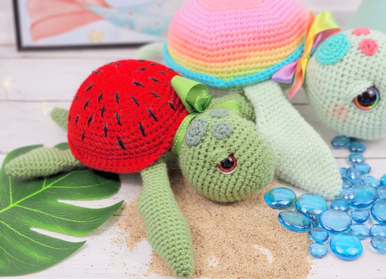 Puede incluir: Dos juguetes de tortuga de crochet, uno con una c&aacute;scara de sand&iacute;a roja y el otro con una c&aacute;scara de arco&iacute;ris. Las tortugas est&aacute;n sobre una superficie blanca con cuentas de vidrio azules.