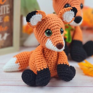 May include: Two crocheted fox plushies, one orange and white with black accents, the other orange with black and white accents. Both are sitting on a white surface.