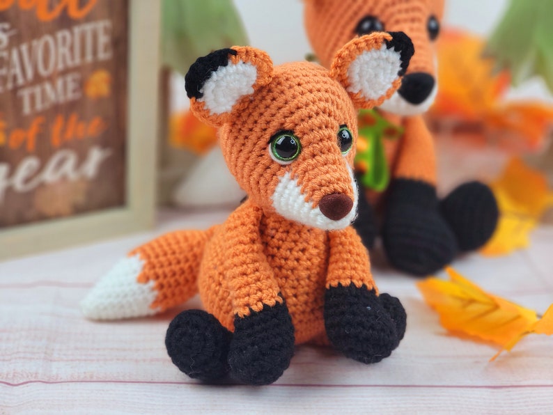 May include: Two crocheted fox plushies, one orange and white with black accents, the other orange with black and white accents. Both are sitting on a white surface.
