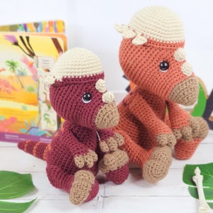 Pu&ograve; includere: Due giocattoli di dinosauro all'uncinetto, uno rosso e uno arancione, con dettagli beige. I dinosauri sono seduti su una superficie bianca con foglie verdi e un osso sullo sfondo.