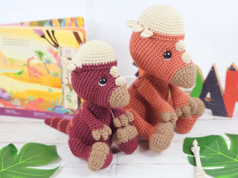 Pu&ograve; includere: Due giocattoli di dinosauro all'uncinetto, uno rosso e uno arancione, con dettagli beige. I dinosauri sono seduti su una superficie bianca con foglie verdi e un osso sullo sfondo.