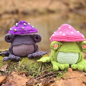 Puede incluir: Dos figuras de rana de ganchillo, una morada y otra verde, cada una con una gorra de hongo. La rana morada tiene un cuerpo morado oscuro y una gorra de hongo morada clara adornada con cuentas moradas. La rana verde tiene un cuerpo verde claro y una gorra de hongo rosa con cuentas blancas.