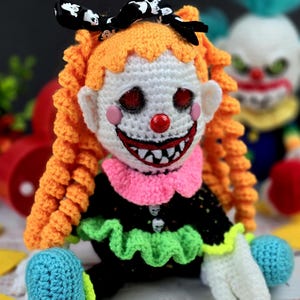 May include: A crocheted clown doll with orange hair, a white face, red lips, and a wide, toothy grin. The doll is wearing a black dress with green and pink ruffles, and has a black bow with white polka dots in its hair.
