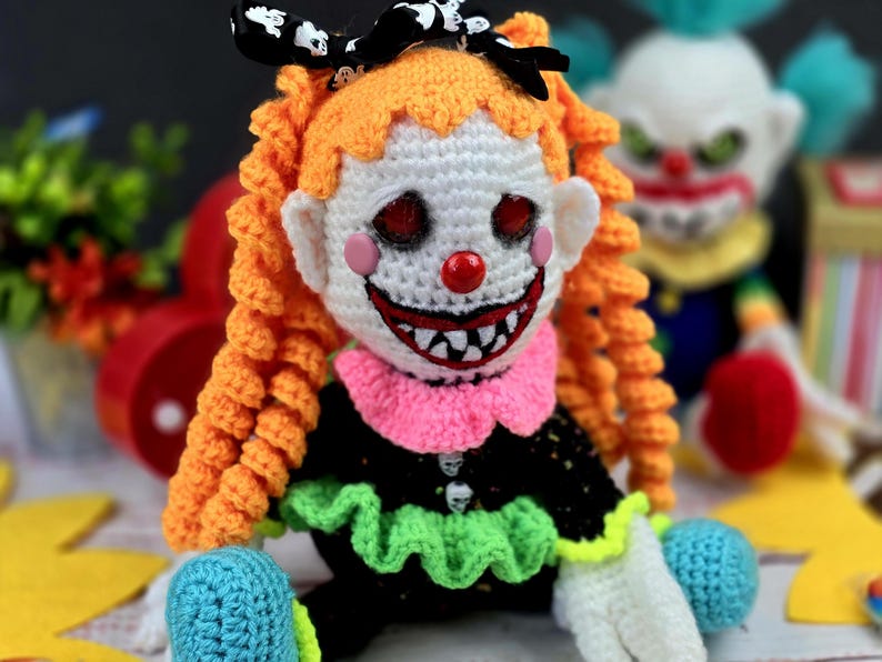 May include: A crocheted clown doll with orange hair, a white face, red lips, and a wide, toothy grin. The doll is wearing a black dress with green and pink ruffles, and has a black bow with white polka dots in its hair.