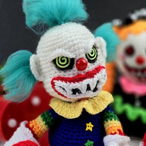 May include: A crocheted clown doll with a white face, red nose, and green eyes. The clown is wearing a rainbow striped shirt and a blue hat with a star.