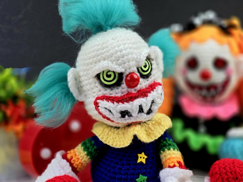 May include: A crocheted clown doll with a white face, red nose, and green eyes. The clown is wearing a rainbow striped shirt and a blue hat with a star.