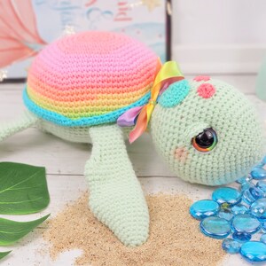 Puede incluir: Un juguete de tortuga de crochet arco&iacute;ris con una concha rosa, un cuerpo verde y grandes ojos negros. La tortuga est&aacute; tumbada sobre una superficie blanca con arena y cuentas de vidrio azules.
