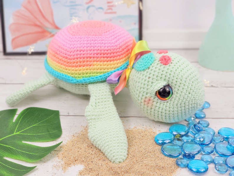 Puede incluir: Un juguete de tortuga de crochet arco&iacute;ris con una concha rosa, un cuerpo verde y grandes ojos negros. La tortuga est&aacute; tumbada sobre una superficie blanca con arena y cuentas de vidrio azules.
