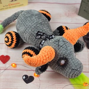 Peut inclure: Un dragon gris crochet&eacute; avec des cornes oranges, des pattes ray&eacute;es noires et oranges et une pointe de queue verte. Le dragon a un n&oelig;ud noir avec des accents de cr&acirc;ne et un pentagramme sur la t&ecirc;te. D&eacute;cor sur le th&egrave;me d'Halloween.