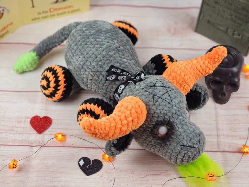 Peut inclure: Un dragon gris crochet&eacute; avec des cornes oranges, des pattes ray&eacute;es noires et oranges et une pointe de queue verte. Le dragon a un n&oelig;ud noir avec des accents de cr&acirc;ne et un pentagramme sur la t&ecirc;te. D&eacute;cor sur le th&egrave;me d'Halloween.