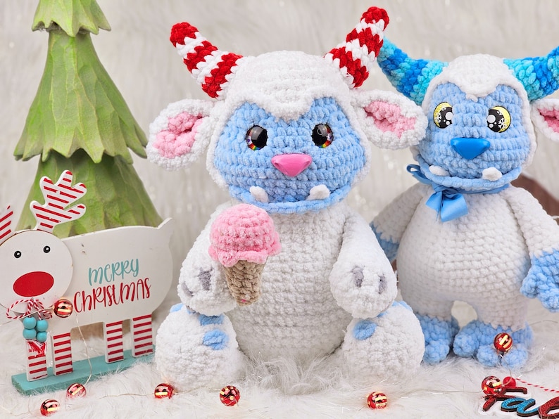 Pu&ograve; includere: Due giocattoli di peluche all'uncinetto, uno blu e bianco con un cono gelato rosa, l'altro blu e bianco con un fiocco blu, davanti a un cartello di renna di legno che dice "buon natale".