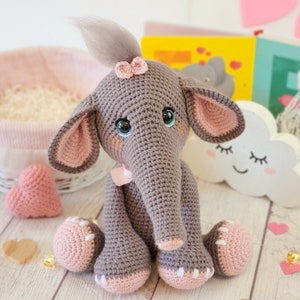 May include: A crocheted gray elephant toy with pink ears, a pink bow, and pink feet. The elephant has blue eyes and a gray yarn tail. The toy is sitting on a wooden surface with pink hearts.