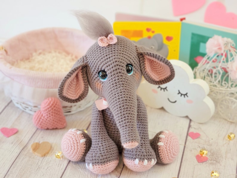 May include: A crocheted gray elephant toy with pink ears, a pink bow, and pink feet. The elephant has blue eyes and a gray yarn tail. The toy is sitting on a wooden surface with pink hearts.