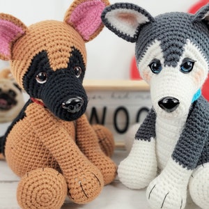 May include: Two crocheted stuffed dog toys. One is a brown and black German Shepherd with pink ears. The other is a gray and white Husky with blue eyes. Both dogs are sitting on a white surface.