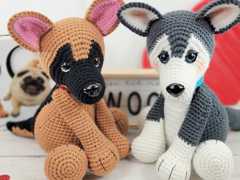May include: Two crocheted stuffed dog toys. One is a brown and black German Shepherd with pink ears. The other is a gray and white Husky with blue eyes. Both dogs are sitting on a white surface.