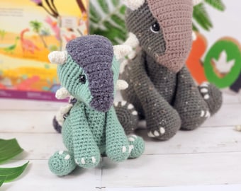May include: Two crocheted dinosaur toys, one green and one brown, sitting on a white surface. The green dinosaur has a purple head and white horns. The brown dinosaur has white spots.