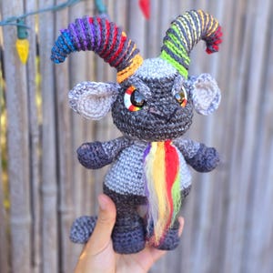 Puede incluir: Un juguete de amigurumi de crochet gris con cuernos arco&iacute;ris y cola. El juguete tiene grandes ojos negros y una expresi&oacute;n gru&ntilde;ona.
