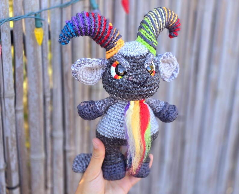 Puede incluir: Un juguete de amigurumi de crochet gris con cuernos arco&iacute;ris y cola. El juguete tiene grandes ojos negros y una expresi&oacute;n gru&ntilde;ona.
