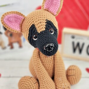 May include: A crocheted brown and black dog with pink ears. The dog is sitting with its front paws tucked in and its back paws extended.