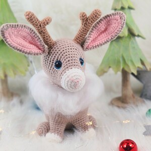 May include: A crocheted reindeer with large blue eyes, brown antlers, and a white fluffy collar. The reindeer is sitting on a white surface, with green Christmas trees and a star in the background. A red ornament is in the foreground.