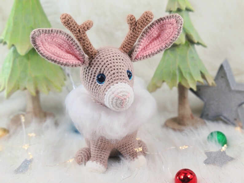 May include: A crocheted reindeer with large blue eyes, brown antlers, and a white fluffy collar. The reindeer is sitting on a white surface, with green Christmas trees and a star in the background. A red ornament is in the foreground.